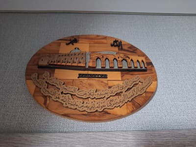 Here's a description of the image:
The image shows a wooden oval plaque mounted on a gray wall. The plaque depicts a stylized scene of Jerusalem. The Dome of the Rock and the Western Wall appear on the plaque. The word "Allah" is written in Arabic script above the buildings. Below the Jerusalem depiction, there is an inscription in Arabic. The word "JERUSALEM" is written in block letters between the buildings and the inscription. The plaque appears to be made of olive wood.