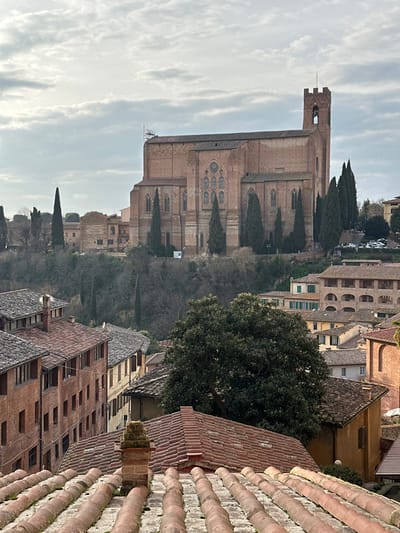 This image captures an elevated, panoramic view of Siena, Italy, under an overcast sky. In the foreground, weathered terracotta tile rooftops with visible moss and a central brick chimney structure descend into the midground. The midground features a dense cluster of historic, multi-story buildings with red brick or pastel-colored facades and dark-tiled roofs, nestled into a sloped landscape. A large, dark green tree with dense foliage occupies the right side of the midground, partially obscuring lower structures.
In the background, positioned prominently on a distant hill, stands the Basilica of San Domenico. This large, imposing brick church exhibits Gothic architectural elements, including tall, arched windows with intricate tracery, buttresses, and a crenellated bell tower on its right side. Scaffolding is visible near the upper left portion of the church's facade, indicating ongoing work. The church is flanked by several tall, slender cypress trees, and a small parking area with multiple white vehicles is discernible to its right. The hilly terrain beneath the church is covered in dense, mostly bare, deciduous vegetation. No discernible individuals or specific human actions are visible within the scene.