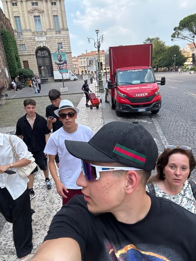 A group of people are posing for a photo in Verona, Italy. A red Iveco truck is parked in the background. One person is pulling a cart with a red crate. Buildings are visible in the background, including one with a poster for "Earth of Glass." Several people are sitting on a stone-paved surface, while others are standing.