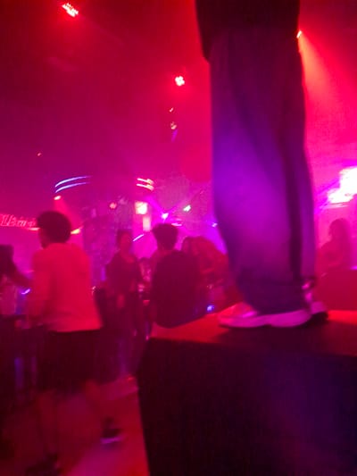 The image depicts a dimly lit nightclub scene, likely in Nanning, China, characterized by vibrant red and purple lighting. In the foreground, a person's legs and feet are visible, clad in dark trousers and white sneakers, standing on a dark, elevated platform. The background is populated by several indistinct figures, suggesting a crowd of people dancing or socializing. Some appear to be in motion, their forms blurred by the low light and the dynamic atmosphere. Neon signs with Chinese characters are faintly discernible in the mid-ground, contributing to the nightclub ambiance. The overall impression is one of a lively, energetic evening event.