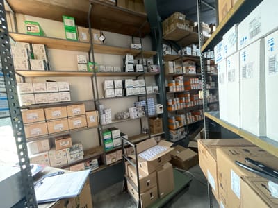 This image captures the interior of a well-stocked warehouse or storage facility, likely located in Campana, Argentina. The scene is dominated by multiple tall metal and wooden shelving units filled with various cardboard boxes and packaged goods, suggesting an active inventory.
The shelves are laden with numerous items, many of which are in white or brown cardboard boxes. Several boxes are explicitly labeled with brand names such as "PHILIPS," "JELUZ," and "FIRLAR." Specific model numbers like "20027" and "40021" are visible on multiple product boxes, indicating a systematic organization of goods. One large box in the upper right section clearly states "GLASS SHELVES," "E-25030," and "MADE IN CHINA," suggesting imported products.
In the foreground, a metal rolling cart is visible, carrying more boxes, some of which are open, revealing smaller packaged items inside. Nearby, a table or desk holds papers and a binder, implying administrative or inventory management tasks are conducted in this area. The floor appears to be concrete, typical of industrial storage spaces.
The lighting is bright and artificial, consistent with an indoor commercial or industrial setting. No people are visible, but the arrangement of goods and the presence of a cart and paperwork suggest ongoing operations of stocking, organizing, or preparing items for distribution. The overall impression is one of a functional and busy storage environment.