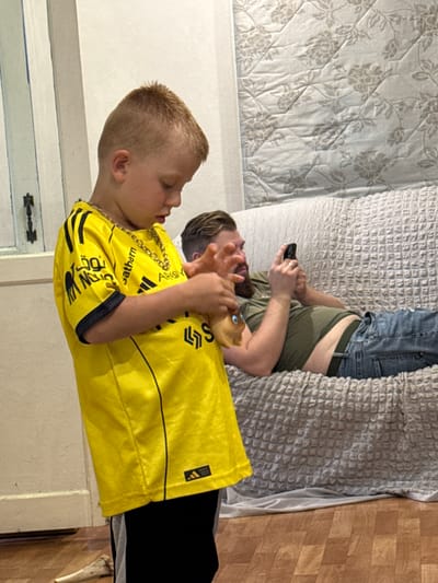 In an indoor setting, a young boy wearing a yellow and black soccer jersey with gold chain necklaces stands in the foreground, looking down at his hands as he manipulates a small, tan-colored object. To his right and in the background, a man with a beard lies on a sofa, holding a smartphone and looking at it. The man is wearing a green t-shirt and blue jeans. The sofa is covered with a textured grey blanket. The environment appears to be a residential interior, with a wooden floor visible in the foreground. The lighting suggests it is daytime, likely indoors, as there is no direct sunlight. The location is identified as the city of Valday, Russia.