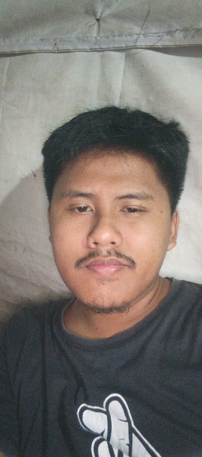 The image is a close-up, selfie-style portrait of a young East Asian male lying down on a light-colored, textured surface, likely a bed or mattress. The man has short, dark hair, prominent eyebrows, dark eyes, and a faint mustache and goatee. He is wearing a dark gray t-shirt with a white graphic design of two hands forming a shape, possibly a heart or peace sign. The background is a textured, off-white material, possibly a sheet or blanket, with a visible seam or fold running horizontally across the upper portion of the image. The lighting appears to be natural and diffused. The image is not clearly associated with the city of Kemuning, Indonesia, or any specific urban environment.