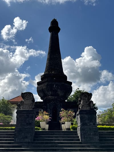 The image shows the Kerta Gosa, a historic building in Klungkung, Bali, Indonesia. The building is a traditional Balinese hall with a tiered roof, featuring intricate carvings and paintings. In the foreground, a stone stairway leads up to the entrance. Flanking the stairway are two guardian statues, known as "Dwara Palas." The sky is blue with scattered white clouds.