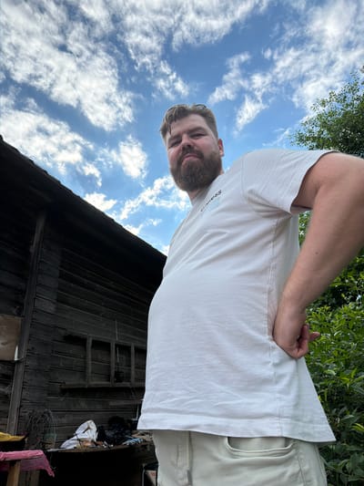 In Бронница, Russia, a man with a beard stands in front of a wooden structure under a cloudy sky. He is wearing a white t-shirt and light-colored pants, with his hand resting on his lower back. The wooden structure appears weathered and has a ladder leaning against it. Various objects, including bags and what looks like wire, are scattered near the base of the structure. Lush green foliage is visible to the right of the man. The perspective of the photo is from a low angle, looking up at the man and the sky.