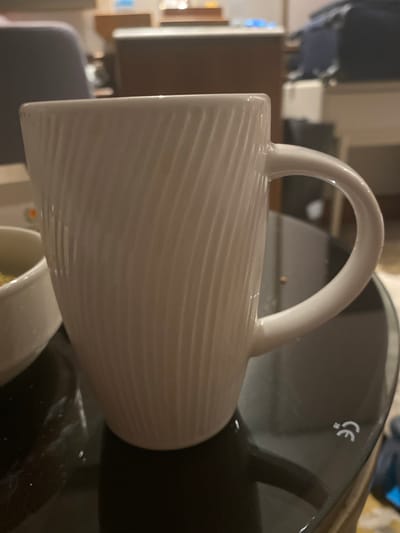 A white mug with a textured pattern is placed on a dark, reflective surface. The mug has a handle and appears to be empty. In the background, out of focus, are pieces of furniture, including a dark chair and a wooden cabinet. There are also some blurred objects on shelves. The overall environment suggests an indoor setting, possibly a living room or dining area. No people are visible in the image. The location in Toroslar, Turkey, is not discernible from the visual information provided in the image.