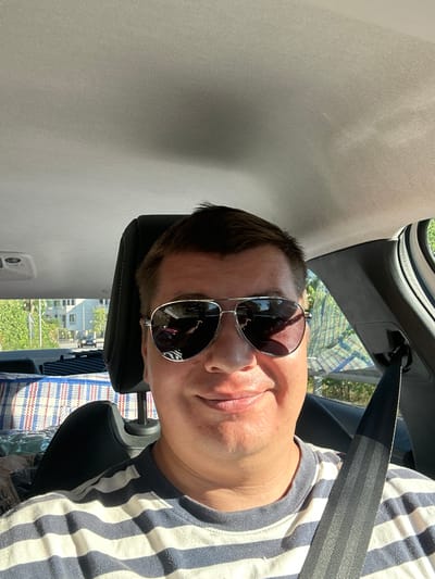 A man wearing aviator sunglasses and a striped shirt is in the passenger seat of a car, looking at the camera. He has short brown hair and is smiling. The car's interior is visible, including the headrest and a seatbelt. Outside the car, trees and buildings can be seen, suggesting an urban or suburban setting. The image is taken from a low angle, looking up at the man.