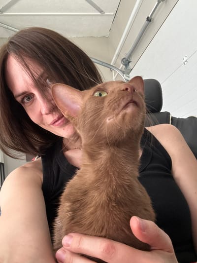 A medium shot captures a woman and her brown cat, likely taken indoors, possibly in a residential garage due to the visible overhead door track and white paneling. The woman, with short brown hair and a black tank top, is looking towards the camera with a slight smile. Her left arm is around the cat, which is held in a cuddled position. The cat, facing upwards and to the right, has green eyes and its mouth is slightly open, giving the impression it is meowing or vocalizing. The background is out of focus, but suggests an industrial or utilitarian space. The scene appears to be a candid moment between the woman and her pet, possibly a selfie. The image does not contain information about the specific location in Konstantinovo, Russia.