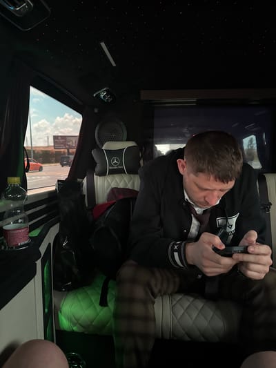 The image shows a man sitting inside a luxury vehicle, likely a Mercedes-Benz van, as indicated by the headrest. He is wearing a dark jacket over a white shirt and a tie, and plaid pants. The man is looking down at his phone, which he is holding in his hands. The interior of the vehicle features a quilted white seat and green ambient lighting. The ceiling is black with small red lights, resembling a starry sky. Through the window, a street scene is visible with cars and a billboard, suggesting an urban or semi-urban environment. The location is identified as Arnavutköy, Turkey.