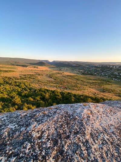 This image captures a panoramic view from a rocky outcrop overlooking a rural landscape in the Krasnomakskoye rural settlement, Ukraine. The foreground is dominated by a large, weathered rock formation, textured with lichen in shades of gray, white, and orange, catching the warm light of the setting sun.
Beyond the rock, the landscape unfolds into a valley characterized by rolling hills covered in dense green vegetation and cultivated fields. The fields are a mix of golden hues from dry grass and patches of green crops, possibly vineyards, given the appearance of ordered rows. A dirt road snakes through the terrain.
In the middle ground, a small village is visible, with a cluster of houses featuring red roofs, interspersed with trees. The village is nestled within a valley, with the surrounding hills forming a natural amphitheater. The light of the setting sun casts long shadows and illuminates the landscape with a golden glow, highlighting the textures and contours of the land.
The background features distant mountains, their silhouettes softened by the haze of distance and the warm light. The sky above is a clear, gradient blue, transitioning from a pale, almost white hue near the horizon to a deep, rich blue at the zenith, indicating either sunrise or sunset. The overall impression is one of serene rural beauty bathed in the golden hour light.