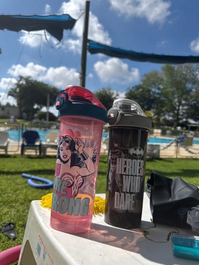 Two character-themed water bottles, one pink with Wonder Woman and text reading "YOU ARE STRONG," and the other dark gray with Batman and text reading "HEROES WHO DARE," are placed on a white plastic table in an outdoor setting. The background features a swimming pool, lounge chairs, and trees under a blue sky with scattered clouds, consistent with a sunny day in Beit Yitzhak-Sha'ar Hefer, Israel. The scene suggests a recreational area, possibly a poolside, where the water bottles are left unattended.