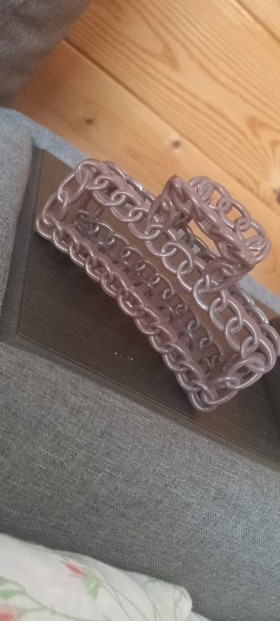 A close-up, overhead shot focuses on a chunky, light purple plastic chain bracelet. The bracelet is laid out on a textured, dark grey fabric surface, which appears to be part of a chair or sofa. Behind the bracelet, a darker brown surface, possibly a book or a tray, is visible. The background consists of warm-toned wooden planks, likely a floor, with visible grain patterns and knots. The image is slightly blurred, with the focus sharpest on the bracelet. There are no people or discernible actions taking place. The setting appears to be indoors, likely in a residential environment. The location in the city of Tersky, Russia cannot be determined from the visual information provided.