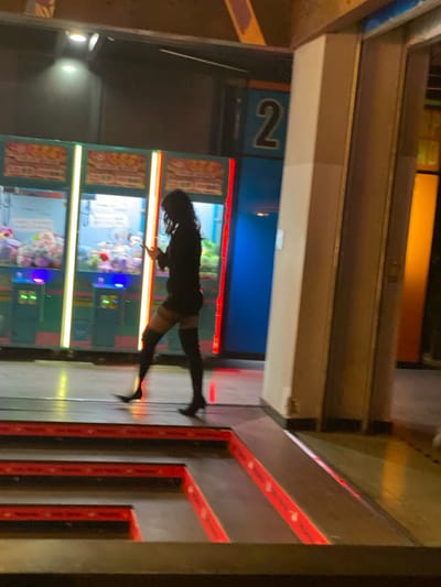 A person stands in front of several arcade claw machines. The machines display various plush toys. The individual is wearing a dark outfit with thigh-high boots. A blue wall is behind the claw machines, and the number "2" is visible above them. To the right of the individual is a gray wall with red accents. Overhead, there are several horizontal structures.
