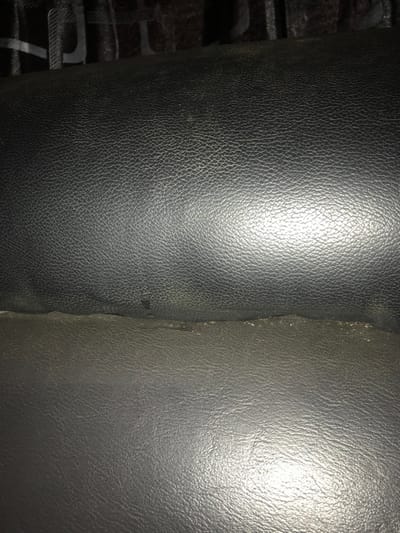 The scene is observed in Oba-Ile, Nigeria. The image depicts a close-up view of two distinct sections of dark, textured material, consistent with synthetic leather or vinyl upholstery, likely comprising parts of a seat or furniture. The upper section displays a fine pebbled grain and exhibits reflections from an ambient or artificial light source. A horizontal seam or crevice, showing minor wear and discoloration, separates this upper section from a lower section of similarly textured material. The lower section also reflects light, indicating a smooth yet textured surface. The background directly above the upper material is dark, out of focus, and shows faint, indistinct patterns, possibly a grille or woven fabric. No individuals, discernible objects other than the upholstery, or specific actions are present within the frame. The immediate environment is an interior space with visible lighting.