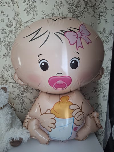 A full shot shows a large, inflatable baby balloon with a pink bow on its head, a pink pacifier, and a yellow baby bottle depicted on its stomach. The balloon is light brown and has large black eyes and blush marks on its cheeks. It is positioned on a white surface against a floral wallpaper with a black and white pattern. To the left of the balloon, a portion of a white fluffy stuffed animal is visible. The scene appears to be an indoor setting, possibly a room decorated for a baby shower or celebration. The exact location in the city of Новобурейский, Russia, cannot be determined from the image alone.