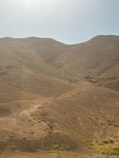The media file depicts a mountainous, arid landscape under a clear, bright sky. The terrain is characterized by steep, eroded slopes covered in dry, brown earth and sparse, green vegetation. The texture of the mountainside is rough and uneven, with visible striations and small depressions. There are subtle variations in color, ranging from light tan to deeper earthy browns, with patches of muted green from the hardy plants clinging to the slopes.
On the left side of the image, a bright, hazy sky dominates, suggesting either early morning or late afternoon light, or possibly atmospheric haze. Several thin, dark lines are visible in the sky, extending from the bottom left towards the upper right, which appear to be power lines or cables, suggesting human infrastructure within this natural setting.
The overall impression is one of a remote, rugged, and somewhat desolate natural environment, likely in a desert or semi-arid region. The sunlight highlights the contours of the mountains, creating shadows that emphasize their ruggedness. There is no discernible activity taking place in the image; it is a still, scenic view of the landscape. The location context of Tamar Regional Council in Israel aligns with the arid, mountainous terrain visible. There is no visible text in the image.