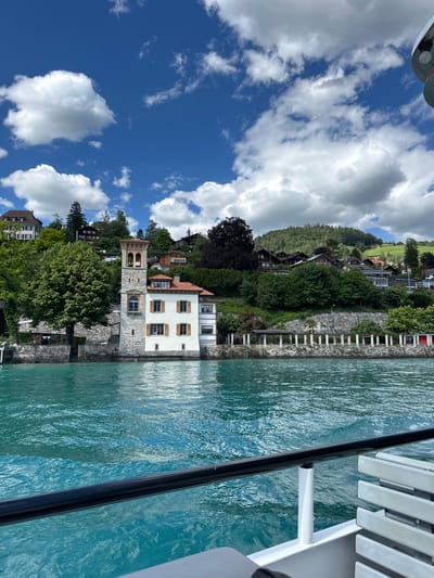 This is a full shot of the picturesque town of Oberhofen am Thunersee, Switzerland, captured from the perspective of a boat on the turquoise waters of Lake Thun. The scene unfolds under a bright blue sky dotted with fluffy white clouds.
Dominating the left side of the frame is a striking white building with a prominent stone tower, featuring arched openings and a terracotta roof. This building appears to be situated on the lakeshore, with a stone wall and lush greenery surrounding it. Several other houses and structures are visible further up the hillside, nestled amongst dense trees and vegetation, suggesting a residential area on the slopes. The architecture of the houses is varied, with some featuring traditional wooden elements.
The water in the foreground is a vibrant blue-green, reflecting the sky and the surrounding scenery. Ripples on the water's surface indicate gentle movement. The edge of the boat on which the photograph is taken is visible in the lower right corner, with a black railing and white seating.
The overall impression is one of serene natural beauty and charming lakeside architecture. The image captures a quintessential Swiss lakeside town on a clear, sunny day.