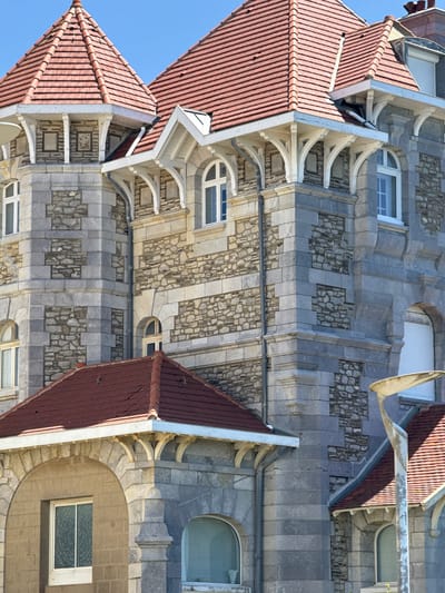 The image shows a close-up of a section of a historic building in Biarritz, France, characterized by its stone masonry and red tile roofs. The architectural style appears to be a blend of traditional European influences, possibly indicative of late 19th or early 20th-century construction. The building's facade is constructed from large, grey stone blocks, interspersed with sections of rougher, smaller stones, creating a textured appearance. Decorative white architectural elements, such as corbels and trim, are visible beneath the eaves of the prominent red-tiled roofs. Several windows are present, some arched and others rectangular, with white frames. The sky in the background is clear and blue. The photograph captures a static, architectural scene, with no people or specific actions depicted. The details suggest a well-maintained but aged structure, typical of the seaside architecture found in the Basque Country.