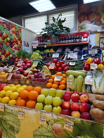 This is a medium shot, eye-level, slightly upward-angled photograph of a produce stall in what appears to be an indoor market, possibly in Orekhovo-Zuyevo, Russia. The stall is overflowing with a vibrant display of fresh fruits and vegetables, artfully arranged on tiered shelves and in baskets.
Dominating the foreground and mid-ground are various fruits, including bright yellow lemons, juicy oranges, and a variety of red and yellow apples. Behind these, plums and other red fruits are visible. Further back, a selection of vegetables is displayed, including green cucumbers, bell peppers in red and yellow, and large, light green zucchini. A head of napa cabbage, wrapped in plastic, is prominently placed on the right side of the stall.
Various handwritten price tags, displaying Cyrillic text and numbers, are attached to the produce, indicating prices such as "244", "835", "1983", "475", "330", and "162". The background reveals more of the market setting, with shelves stocking bottled beverages, a decorative fruit mural on the wall, and overhead fluorescent lighting. The overall impression is one of abundance and freshness, typical of a bustling local market. No people are clearly visible in the frame. The scene is well-lit, suggesting it's likely daytime.