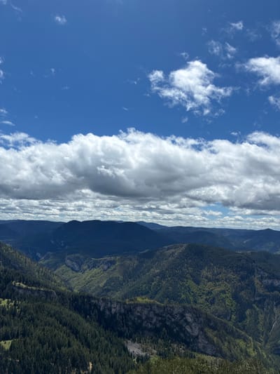 The image shows a landscape view with a blue sky, clouds, mountains, and forest. The sky occupies the left side of the image, featuring scattered white clouds. A thick band of white clouds stretches vertically down the center of the image. The mountains occupy the right side, with varying shades of green indicating forest cover. The location is likely Giovren, Bulgaria. There are no people visible in the image.