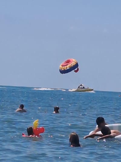 The image depicts a bright, sunny day at the sea, likely in Dzhubga, Russia, as indicated by the context. The water is a vibrant blue, with small waves indicating activity. In the distance, a boat is moving across the water, leaving a white wake. Above the boat, a large, circular inflatable, possibly a parasail, is suspended in the air, featuring red, yellow, and blue sections.
Closer to the foreground, several people are visible in the water. To the left, a person is floating, with their back to the camera. In the center left, a red inflatable toy, resembling a character with yellow wings and black ears, is floating. Further to the right, two children are in the water near an inflatable raft. One child, with dark hair, is looking towards the camera, while another, partially obscured, is also in the water. In the far right, two individuals are on an inflatable raft, with one person partially submerged. The overall scene is one of leisure and recreation on a summer day at the beach.