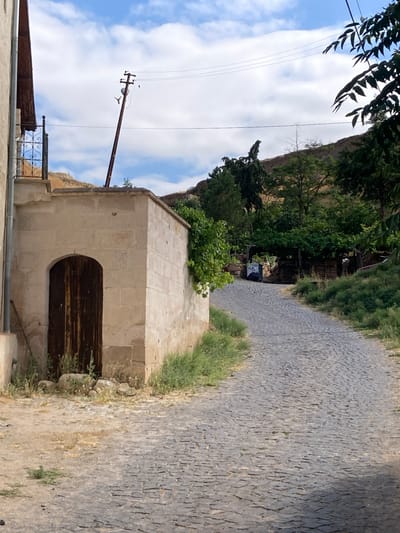 The image depicts a view of a narrow, winding cobblestone street in Avanos İlçe Merkezi, Turkey. The street is flanked by buildings constructed from light-colored stone. To the left, a building with a vertical downspout is partially visible, adjacent to a stone structure with a dark wooden arched doorway. This doorway is set into a light beige stone wall, with some patchy green vegetation growing around its base.
The cobblestone street itself is made up of irregular, gray stones, with some grass growing in the cracks. The street curves to the right and leads uphill. On the right side of the street, the terrain rises and is covered in dense green vegetation and trees.
In the background, a utility pole with wires is visible against a bright blue sky with scattered white clouds. The pole is leaning at an angle. Further up the hill, more trees and rocky terrain are present. A dark, possibly metal bin with some writing or symbols on it is situated near the base of the uphill vegetation. The overall scene suggests a historical or rural setting within the town.