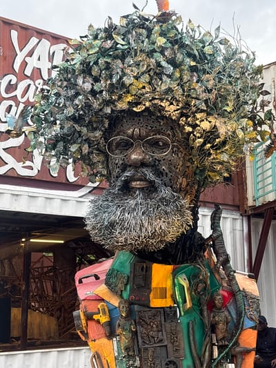 This image depicts a large, intricate sculpture of a man, likely an artistic representation, situated outdoors. The sculpture features a metallic face with round glasses and a bushy, wire-like beard. Its head is adorned with a dense canopy of metallic leaves and branches, giving it a tree-like appearance. The torso of the sculpture is constructed from a collage of various found objects and materials, including what appears to be car parts, tools, circuit boards, and smaller figurines or carved elements. One notable element on the torso is a yellow power drill. The sculpture is positioned in front of a weathered, dark red shipping container with large white lettering.
The overall scene suggests an outdoor art installation or exhibition. The backdrop includes additional shipping containers and some structural elements, hinting at an urban or industrial setting. The lighting indicates it is daytime, likely overcast given the diffused light. The presence of a person in the lower right corner, wearing a dark cap and jacket and sitting down, suggests this may be a public space, possibly an art park or a venue displaying this work. The location is identified as the city of Karum, Nigeria.