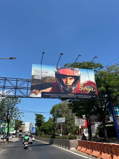 This is a street-level, wide-angle shot capturing a billboard advertisement prominently displayed against a clear blue sky. The billboard features a close-up of a man wearing a red motorcycle helmet, with text that translates to "Effort does not betray results." Below this, smaller text reads "WARNING: Because I smoke, I am experiencing throat cancer. Quit smoking service (0800-177-6565)." Another line states, "NOT FOR SALE AND GIVING TO PEOPLE UNDER 21 YEARS OLD AND PREGNANT WOMEN." The bottom right corner of the billboard has a logo that says "Kita adalah Surya," which means "We are Surya."

The billboard is supported by a large metal scaffolding structure. In the foreground, a street scene unfolds with vehicles, including a scooter with a rider in a green jacket, and cars driving on an asphalt road. Trees line the side of the road, and various buildings and other billboards are visible in the background. The overall impression is a bustling urban environment under a bright, sunny sky. The image was taken in Makassar, Indonesia.