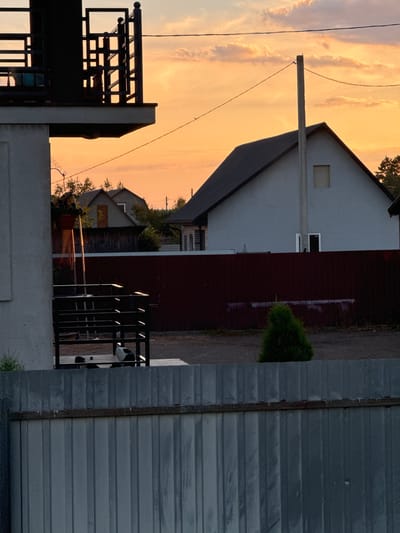 In Chemerne, Ukraine, at dusk, a residential scene unfolds. A light-colored building with a dark roof dominates the middle ground, a concrete utility pole stands in front of it, and a single, small, dark-colored window is visible on the upper portion of the building. A maroon metal fence runs horizontally across the mid-section of the image, with a small, green, cone-shaped bush in front of it. In the foreground, a tall, corrugated metal fence obscures the lower part of the scene. To the left, a portion of a light-colored building and a dark metal railing are visible, suggesting a balcony or porch. The sky is filled with orange and pink clouds, indicative of sunset.