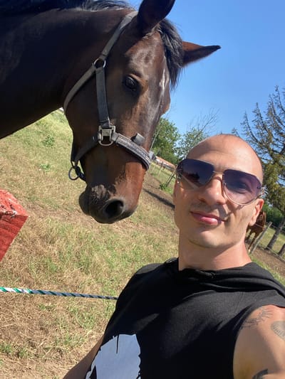 The image captures an outdoor scene in Slavovitsa, Bulgaria, featuring an adult male and a horse. An adult male, bald, with dark aviator sunglasses and a black sleeveless hooded garment, occupies the lower right and center of the frame, taking a selfie. A tattoo is visible on his right upper arm. A dark brown horse wearing a dark halter with a metal ring and buckle is positioned in the upper left and central areas, its head close to the man. Its right eye, nostril, and part of its mane are visible. The ground is covered with dry, patchy grass and some exposed earth. A red rectangular object is partially visible on the far left foreground. A green and white striped rope lies horizontally across the lower left ground. The background consists of a clear blue sky, scattered deciduous trees, and several tall coniferous trees on the right. A fence line and another horse are visible in the mid-ground behind the man.