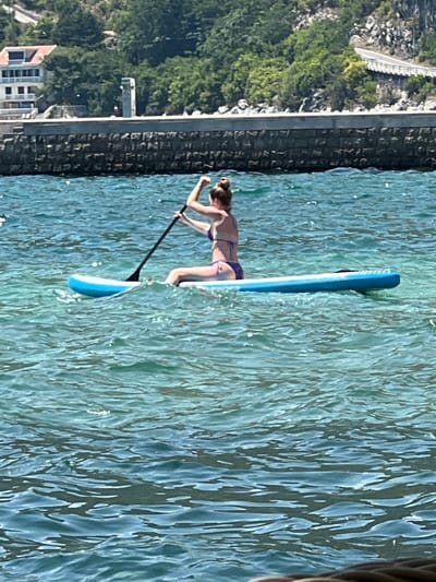 A woman is paddleboarding on the turquoise waters of the Bay of Kotor in Risan, Montenegro. She is wearing a purple bikini and is seated on a blue paddleboard, holding a black paddle. The paddleboard is in motion, creating small ripples on the water's surface. In the background, a stone embankment lines the shore, topped with trees and a building. The water is clear and reflects the bright sunlight.