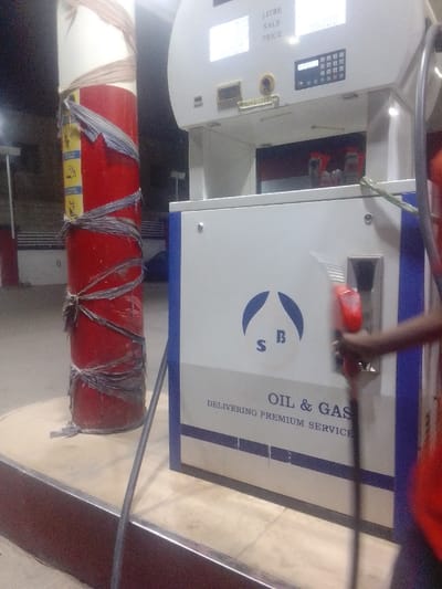 A red cylindrical post wrapped in gray material stands next to a gas pump. The pump features a digital display showing "LITRE SALE PRICE." It also has a logo with "S B" beneath a stylized droplet, and the text "OIL & GAS DELIVERING PREMIUM SERVICE." A person's arm holding a red gas nozzle is partially visible on the right side of the frame. The scene appears to be at a gas station during the evening.