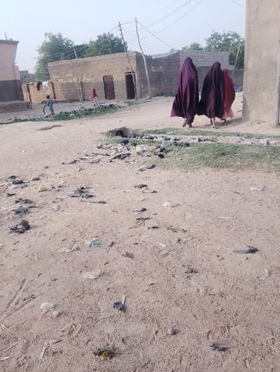 This image captures a street scene in Chiranchi, Nigeria, under daylight conditions, likely late morning or afternoon given the soft lighting. The setting appears to be a rural or semi-urban area characterized by unpaved, sandy ground serving as a pathway.
In the mid-ground, three individuals, likely women, walk away from the viewer. Two are dressed in dark purple, flowing burqas or hijabs that completely cover their faces and bodies, while the third, walking beside them, appears to be wearing a reddish garment beneath a partially visible dark covering.
The background features simple, single-story structures built from concrete blocks, some showing signs of wear or being unfinished. One building on the left has laundry hanging outside. Utility poles with overhead wires crisscross the sky, suggesting basic infrastructure. Two children are visible further back; one child in light blue clothing is playing with a hoop-like object, while another child in red stands near a building entrance.
The foreground and mid-ground of the sandy path are extensively littered with various debris, including plastic bags, wrappers, and other small pieces of trash, indicating a lack of formal waste disposal. A small, open culvert or drainage pipe is visible embedded in the ground to the right of the path. Sparse patches of green vegetation grow along the edges of the path. The overall impression is one of everyday life in a community with limited resources, focusing on the movement of people and the surrounding environment. No discernible text is visible in the image.