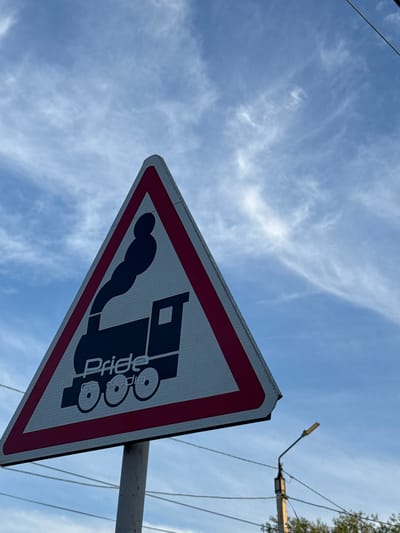 A triangular road sign with a red border is positioned against a blue sky with wispy white clouds. The sign features a black silhouette of a steam train with smoke billowing from its chimney. Below the train, the word "Pride" is visible in white text, followed by what appears to be a stylized representation of train wheels. The sign is mounted on a grey metal pole. In the background, a streetlight and power lines are visible, along with the tops of green trees. The image was taken in the city of Petropavl, Kazakhstan.