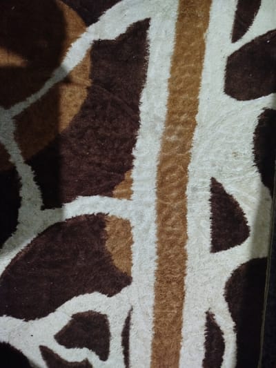 A close-up shows a textured rug or mat with a pattern resembling animal markings, potentially giraffe or abstract spots. Dark brown blotches are contrasted by lighter beige and white shapes. The texture appears plush and possibly soft, with a noticeable pile. The composition suggests only a section of a larger patterned surface. The lighting is uneven, causing variations in shadow and depth.