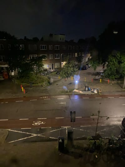 The image depicts an outdoor scene at night, likely in an urban environment, possibly Utrecht, Netherlands, given the presence of bicycles and brick buildings. The foreground shows a wet asphalt road with a red-colored bicycle lane marked with white lines, including a bicycle symbol. A pedestrian crossing with white diagonal markings is also visible. A dark trash can is positioned near the edge of the road.
In the middle ground, a paved area with trees and bushes is present. Several streetlights are visible, one emitting a blueish glow. A collection of trash and debris is piled near a bin. A single white bicycle is parked in this area. Numerous other bicycles are parked further back, leaning against a brick building. This building appears to be residential, with multiple windows illuminated. Yellow and red bollards are placed along the edge of the paved area.
The background shows more brick buildings and trees, silhouetted against a dark night sky. The overall impression is a somewhat neglected urban space at night, with evidence of both residential activity and public amenities.