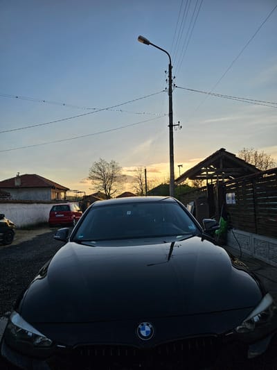 The image captures a tranquil street scene at either sunrise or sunset, characterized by a warm, golden glow on the horizon and a clear, blue sky overhead. The setting is a residential street, likely in a rural or suburban area of Krasnovo, Bulgaria.
In the foreground, a dark-colored BMW SUV is parked on the right, its shiny hood and windshield reflecting the surrounding sky and a nearby utility pole. The distinctive BMW logo is clearly visible on the vehicle's grille. To the left, a tall, black utility pole equipped with a street light and numerous electrical wires stands prominently, with wires stretching across the sky.
Mid-ground details include an asphalt road surface that appears slightly reflective. Further down the street, a red hatchback car is parked, and its Bulgarian license plate, "EB 0894 CA," is partially visible. In the background, various buildings are seen, including a wooden structure with a sloped roof behind the BMW, and a house with a red tiled roof further down the street. Bare-branched trees silhouette against the bright horizon. There is no discernible activity or any people present, contributing to a serene atmosphere.
