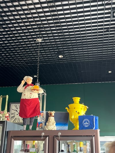 This image captures a display on a shelf, likely in a shop or restaurant, in the village of Akhlebaikha (Kovriginskii S/S), Russia. The backdrop is a dark green wall. Dominating the left side of the display is a life-sized mannequin of a chef, wearing a white chef's coat, a red apron, and a red neckerchief. The chef holds a transparent container with what appears to be cheese or food samples. To the right of the chef, a large, bright yellow samovar is prominently displayed. Next to the samovar is a blue fabric bag with a circular white logo featuring a building and text. Behind the chef, a large wall clock with Roman numerals is visible. To the right of the clock and slightly in front of it are two framed pictures of men in astronaut suits. A small, white decorative object resembling a cow or bull, adorned with a bell, is positioned between the astronaut pictures and the samovar. On the shelf below, various bottles of alcohol are visible through the glass doors of a cabinet. The ceiling above features a black grid with recessed lighting. The overall scene appears to be a themed display, possibly related to culinary arts or Russian culture, set within a commercial establishment.