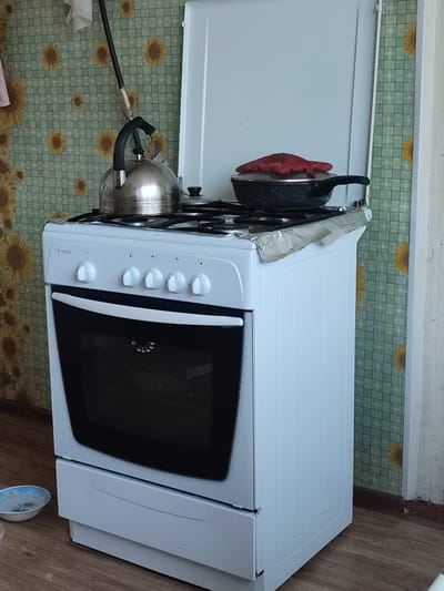 A white gas stove with four burners and an oven is shown in a domestic setting. A silver kettle sits on one of the burners, and a black frying pan with a red lid is on another. The stove is placed in front of a wall covered in green checkered wallpaper with a sunflower pattern. The floor is made of wooden planks. To the left of the stove, a small blue and white bowl is visible on the floor. The scene appears to be indoors, likely a kitchen, given the presence of the stove. The lighting suggests it is daytime. No people are visible in the image. The brand name "GEFEST" is visible on the front of the stove, above the oven door.