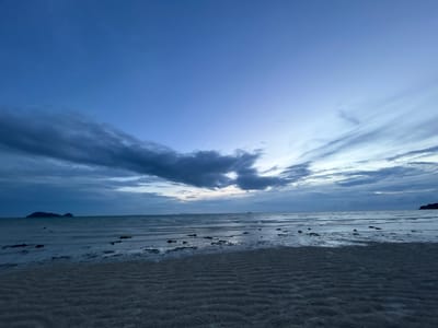 This image captures a serene beach scene at dusk in Ko Pha-ngan District, Thailand. The foreground is dominated by rippled sand, indicating the receding tide. The middle ground features the calm ocean, with shallow water reflecting the twilight sky and scattered dark objects, likely seaweed or rocks, visible in the water. In the distance, a silhouette of islands can be seen on the horizon, adding depth to the seascape. The sky transitions from a deep blue at the top to lighter shades of blue and purple near the horizon, with scattered dark clouds creating dramatic streaks across the sky. The overall mood is peaceful and tranquil, characteristic of a quiet evening by the sea. The time of day is clearly dusk or twilight, indicated by the soft, diffused light and the deep blue hues of the sky. There is no visible text in the image.