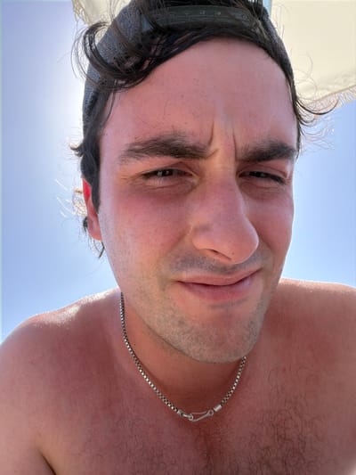 A close-up portrait of a man with dark hair and eyes, taken from a low angle, suggests he's holding the camera above him. He's wearing a gray cap and a silver chain around his neck. The background is a bright blue sky with a hint of sunlight. The man has fair skin and appears to have some stubble. His expression is a slight smirk.