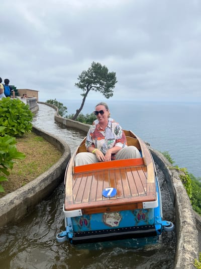 A woman is riding in a wooden boat on a water flume ride. The boat is decorated with blue panels featuring a tiger and a butterfly. A blue circular number "7" is visible on the wooden deck of the boat. The woman is smiling and wearing sunglasses and a colorful shirt. She is seated in the boat with her hands resting on her lap. The water flume follows a winding path alongside a grassy embankment and a concrete wall. In the background, the vast expanse of the sea is visible under a cloudy sky. The location is Monte Igueldo, an amusement park in San Sebastián, Spain, known for its vintage attractions and panoramic views of the Bay of Biscay.