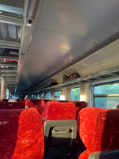 The image shows the interior of a train carriage, likely in Tulovo, Bulgaria. The scene depicts rows of red velvet seats, some of which appear worn. Luggage, including a patterned bag and a black and white checkered bag, is visible on overhead racks. The lighting suggests it is daytime, with sunlight streaming through the windows, casting long shadows. The windows offer a view of greenery outside, hinting at a rural or semi-rural setting. The overall atmosphere is one of travel, with the empty seats and luggage suggesting passengers have either disembarked or are about to board. No people are clearly visible in the image, but reflections in the windows may show glimpses of them. The image is oriented vertically. There is no visible text in the image.