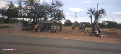 A roadside gathering shows several people under a covered structure on the left, with a small group seated. Further right, more people are present near a tree. Two motorcycles are parked in between the two groups. A road runs along the foreground of the scene. In the background, power lines and greenery are visible. The scene appears to be a roadside stop or small informal market.