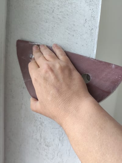 The image shows a close-up view of a person's hand holding a triangular piece of sandpaper against a light-colored, textured wall. The sandpaper is dark reddish-brown and has a hole in the center. The hand is positioned to grip the sandpaper, and its fingers are pressed against the wall, indicating the action of sanding. The wall appears to be a rough plaster or concrete surface. The lighting suggests an indoor setting, likely during the daytime, with no strong shadows. The focus is on the texture of the wall and the action of sanding, with the hand and sandpaper being the most prominent subjects. The background is blurred, with hints of a corner or edge of the wall visible, and a lighter, possibly painted surface. The overall scene depicts a DIY or renovation activity. No text is visible in the image. The provided location context is Ходаево, Russia.