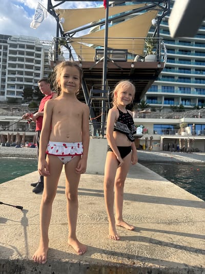 Two young girls are standing on a concrete walkway next to a body of water. The girl in the foreground is wearing white bikini bottoms with strawberries on them. The girl behind her is wearing a black bandana-style bikini top and black bottoms. Both girls are smiling. Behind them, a modern hotel building with many windows is visible. The sky is blue with some clouds. The sun is shining, casting shadows on the walkway. A flag with text on it is partially visible on the left. There is also a person in a red shirt with a fishing rod visible in the background. The scene appears to be taking place in a resort or coastal area, possibly Yalta, Ukraine, during the daytime.