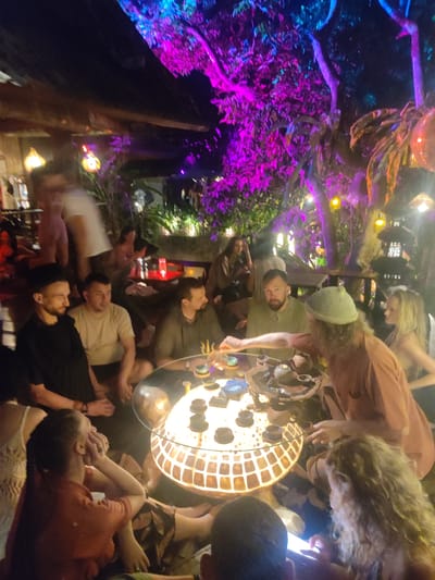 A group of approximately 15 people are gathered around a low, illuminated, round table for a tea ceremony in an outdoor setting, likely a cafe or private yard. The location is Lokaserena, Indonesia, judging by the lantern design and overall tropical atmosphere. The table is set with multiple small teacups, a teapot, and implements for preparing tea. One man, wearing a green beanie and an orange shirt, seems to be leading the ceremony, using a long, thin tool. The other participants are sitting on the ground, some observing the ceremony and others socializing. A woman with curly hair in the foreground is looking at her phone. The background is filled with lush vegetation, brightly lit with pink and blue lights, and decorative lanterns. A building structure with wood beams is partially visible in the background. The atmosphere appears relaxed and informal.