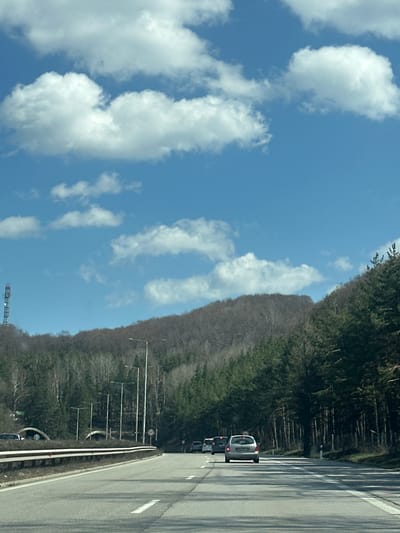 The image depicts a highway scene with a bright blue sky featuring scattered white clouds. A dense forest lines one side of the road, while a few cars are traveling on the paved highway surface. The location cannot be confirmed as Gorna Vasilitsa, Bulgaria, based solely on the image.