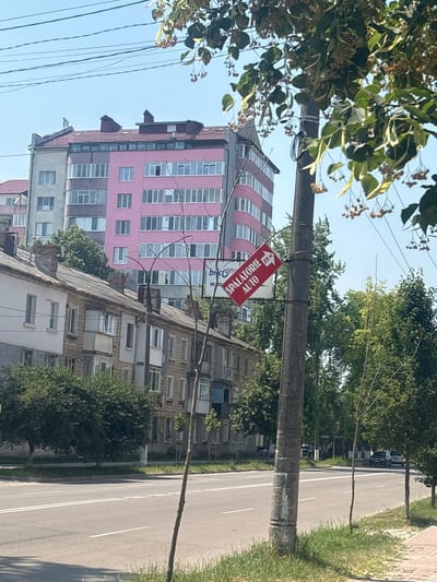 The image depicts an outdoor scene in Ungheni, Moldova, likely during the daytime on a clear day. The perspective is tilted, with the ground appearing on the right side of the frame and the sky on the left.
In the foreground, lush green foliage from a tree with small dried seed pods drapes over the top left of the image. A rough, concrete utility pole stands vertically in the mid-ground, to which a red directional sign is attached. The sign is rectangular with an arrow pointing to the left and contains text in a light color. Visible text on the sign reads "SPALATORIE AUTO" and "70M". This indicates a car wash is located 70 meters to the left.
Behind the utility pole, there are several buildings. The most prominent is a tall, modern building with a pink facade punctuated by rows of white-framed windows. To the right of this, and slightly lower, is an older, multi-story building with a grey facade, also featuring numerous windows and balconies.
Further in the background, a tree-lined street runs parallel to the buildings. Cars are visible parked along the street, suggesting a residential or commercial area. The overall impression is of a typical urban or suburban street scene in Moldova. No people are clearly visible. The lighting suggests it is daytime, and the clear sky indicates fair weather.