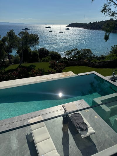 The image depicts a luxurious outdoor setting with a swimming pool overlooking a scenic bay in Vouliagmeni, Greece. The pool, a prominent feature in the foreground, is rectangular and filled with clear turquoise water, reflecting the bright sunlight. Two white sun loungers are positioned on the pool deck, one with a dark gray throw draped over it. The deck itself is paved with large, light gray stone tiles.
Beyond the pool, a vibrant green lawn slopes down towards a line of trees and lush vegetation. In the distance, the shimmering Aegean Sea stretches out, dotted with several white yachts and smaller boats. The coastline is visible, characterized by rocky outcrops and dense greenery. The sky is a clear, bright blue, with a few wispy clouds on the horizon. The sun, high in the sky, casts a strong glare on the water, creating a dazzling effect. The overall impression is one of a serene and upscale vacation destination.